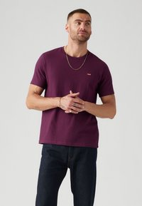Violetinis medvilninis marškinėliai su apvaliu kaklu ir trumpomis rankovėmis, išryškinantis mažą raudoną Levi's logotipą kairėje krūtinės pusėje, derinami su tamsiai mėlynomis džinsomis.