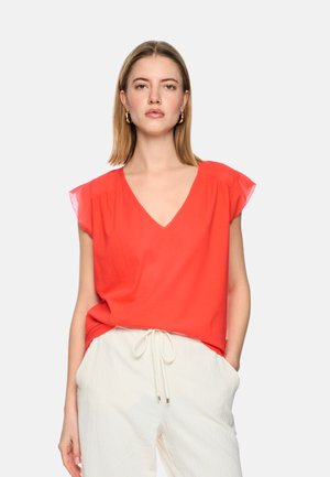 Young woman with shoulder-length blonde hair wearing gold hoop earrings, a bright red V-neck blouse, and white drawstring pants.