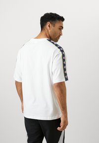 T-shirt en coton blanc à manches courtes, avec une rayure bleu marine sur les épaules et des accents de logo colorés. Vue arrière.