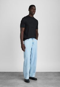 Νεαρός άνδρας φορεμένος με μαύρο κοντό μανίκι πουκάμισο, ανοιχτό μπλε παντελόνι σε φαρδιά γραμμή, και μαύρα παπούτσια, στέκεται με το ένα χέρι στην τσέπη.