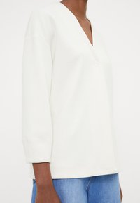 Vit topp med mjuk textur och v-ringning, med knäppning fram och 3/4-långa ärmar. Bärs över ljusblå denim.