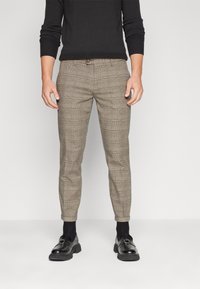 Man draagt slim-fit bruine geruite broek, zwarte sokken, zwarte instappers en een zwart lange mouwen trui, staande op een effen witte achtergrond.