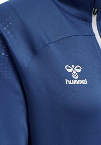 Giacca sportiva blu realizzata in tessuto liscio, con colletto alto, zip bianca e logo Hummel. Presenta dettagli testurizzati sulle spalle.