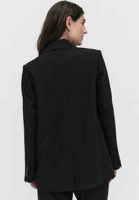 Musta bleiseri, jossa on jäsennelty muotoilu, kärjellinen kaulus ja yksi halkio. Valmistettu sileästä kankaasta, jossa on nappikiinnityksellä varustetut hihat ja istuva leikkaus.