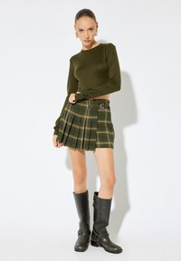Grünes Langarm-Oberteil kombiniert mit einem gefältelten karierten Minirock mit Gürtel. Das Model trägt knielange schwarze Lederstiefel.