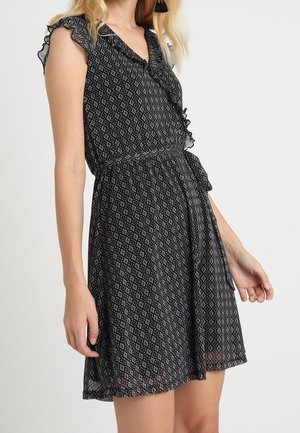 Kvinde iført sort ærmeløs kjole med hvidt diamantmønster, flæseærmer og slå-om-stil bundet i taljen.