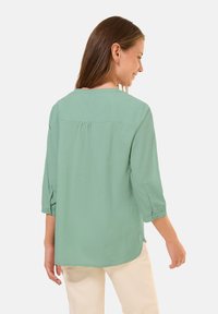 Blusa casual verde chiaro con maniche a tre quarti, dettaglio di piega posteriore e scollo rotondo, realizzata in tessuto liscio.