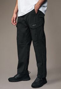 Schwarze Cargo-Hosen mit lockerer Passform, die über mehrere Taschen und eine glatte Textur verfügen. Sie werden mit schwarzen Sportschuhen kombiniert, um einen einheitlichen Look zu erzielen.