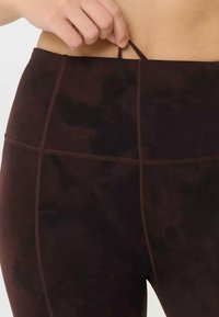 Main tirant une petite boucle de tissu sur un legging taille haute marron foncé avec des lignes de couture visibles.