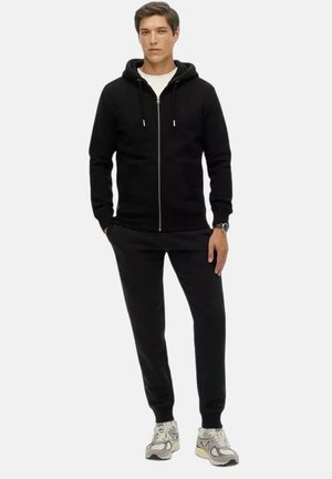 Joven de pie con las manos en los bolsillos, vistiendo una sudadera negra con cremallera, pantalones deportivos negros, camiseta blanca y zapatillas grises sobre un fondo blanco.