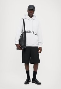 Mann in weißem Hoodie mit Text, schwarzen Shorts, schwarzem Cap, schwarzen Schuhen und mit einer schwarzen Lederschultertasche vor weißem Hintergrund.