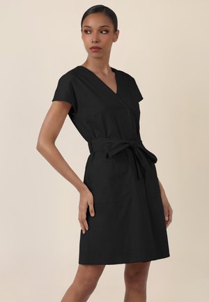 Frau trägt ein schwarz Wickelkleid mit kurzen Ärmeln und Gürtel in der Taille vor einem neutralen beige Hintergrund.