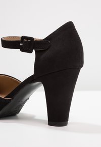 Czarne zamszowe buty na wysokim obcasie z paskiem na kostce i kwadratową klamrą. Okrągły czubek i teksturowana czarna podeszwa. Prosty design z smukłym konturem.