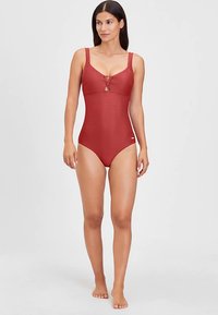 Rode eendelige badpak met geribbelde textuur, verstelbare bandjes, sleutelgatdetail aan de voorkant en bescheiden beenopeningen. Effenkleurig zonder patronen.