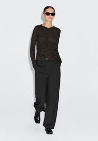 Zwarte top met lange mouwen en geplooide textuur, gecombineerd met losse zwarte broek. Accessoires zijn onder andere donkere zonnebril en glanzende zwarte schoenen.