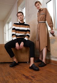 Suéter riscado em preto, branco e castanho; vestido caqui com bolsos; tamancos pretos; chão de madeira; texturas suaves; design minimalista.