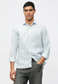 Férfi világos kék csíkos ingben, felgöngyölt ujjakkal és sötét szürke nadrágban, az egyik kezét a zsebében tartva áll egy fehér háttér előtt.