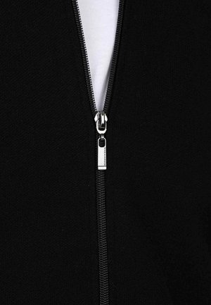 Veste noire zippée présentant une fermeture éclair métallique avec un tirette. Tissu texturé réfléchissant la lumière et une couche intérieure blanche partiellement visible.