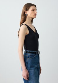 Femme aux longs cheveux bruns, portant un top noir sans manches, un jean bleu, une ceinture noire et des boucles d'oreilles bleues en forme de fleur, debout de profil.
