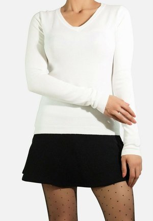 Pull-over blanc à manches longues en V, fabriqué dans un tissu lisse, associé à une mini jupe noire évasée et des collants noirs à pois.