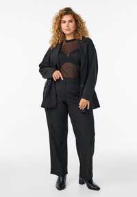 Črn kostim, ki vsebuje krojni blazer in ohlapne hlače. Pod njim je prosojna majica s črnimi pikami. Obut je v črne gležnjarje.
