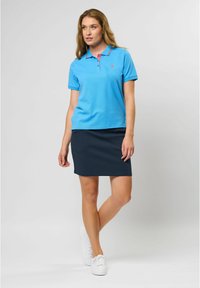Lys blå polo med rosa krage, marineblå skjørt og hvite sneakers. Polotrøyen har korte ermer og en liten logo på brystet.