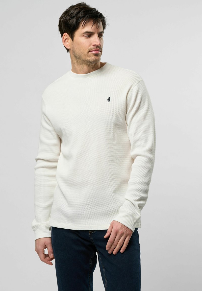 Witte wafelstructuur lange mouwen shirt met geribde manchetten; heeft een klein zwart logo op de borst. Gecombineerd met donkere jeans.