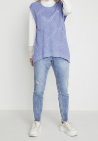 Lichtpaarse gebreide mouwloze trui met een ruitpatroon, gedragen over een witte langemouwen top, gecombineerd met lichtblauwe skinny jeans.