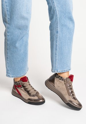 Person trägt hellblaue Jeans und taupefarbene High-Top-Sneaker mit roten und metallischen Akzenten, steht vor einem schlichten weißen Hintergrund.