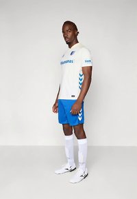 Mužský futbalista stojaci, oblečený v bielom drese s modrými logami sponzorov, modrých šortkách, bielych vysokých ponožkách a bielych kopačkách na jednoduchom pozadí.