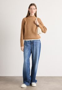Pulover maro cu dungi, gât înalt, asortat cu blugi lejeri albaștri și teniși albi. Modelul ține o geantă mică de culoare crem.