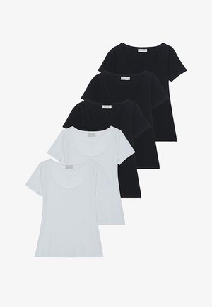 Femme debout devant un fond blanc, portant un haut noir à manches courtes, un pantalon large vert foncé et des chaussures noires tressées.
