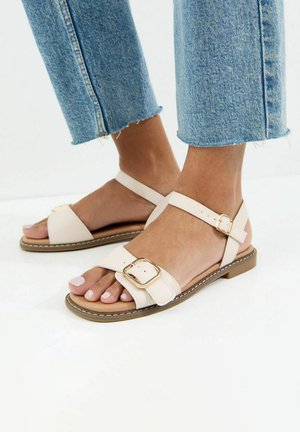 Voeten in lichtbeige platte sandalen met gouden gespen, gecombineerd met cropped lichtblauwe spijkerbroeken tegen een witte achtergrond.