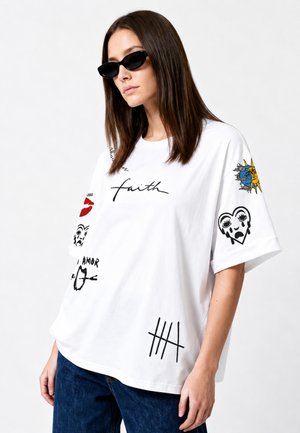 Vrouw met zwarte zonnebril en een wit oversized T-shirt met zwarte en gekleurde grafische prints en blauwe spijkerbroek tegen een effen achtergrond.
