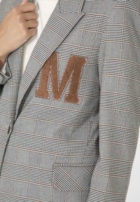Grijze geruite blazer met een bruin vilten "M" embleem, gestructureerd ontwerp, reverskraag en een enkele borstzak.