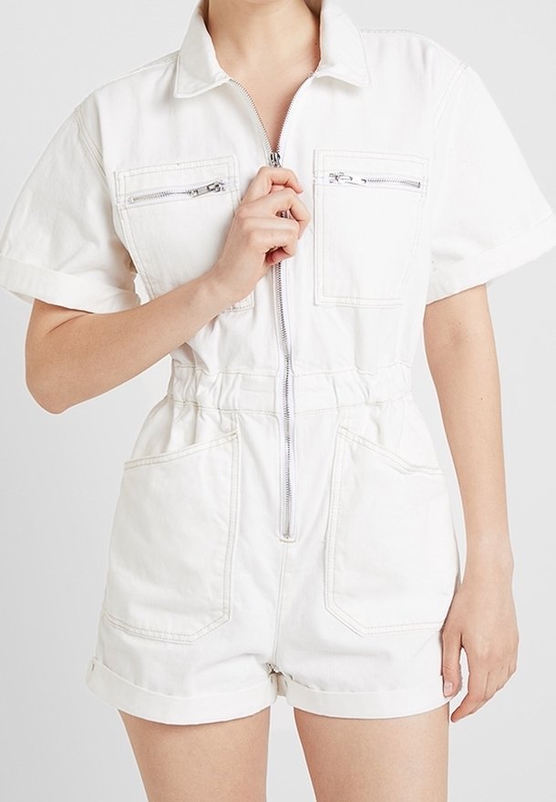 Femme portant une combinaison-short blanche à manches courtes avec fermeture éclair et poches avant, tenant la fermeture éclair au niveau de la poitrine.