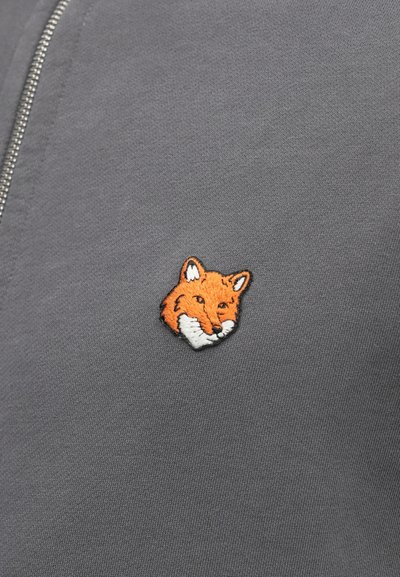 Gray fabric with a smooth texture featuring a small, embroidered fox head in orange and white, located on the left side near the zipper.