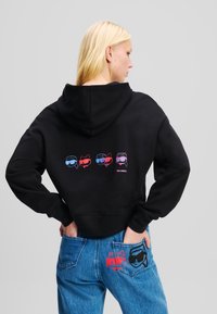 Čierny skrátený teplákový sveter s kapucňou, na zadnej strane s farebnými grafikami štylizovaných postáv. V páre so modrými džínsami s ozdobnými prvkami.