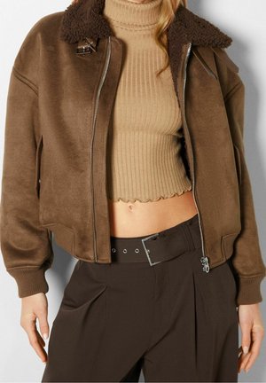 Vrouw draagt een cropped bruine shearling jas over een beige geribde coltrui crop top en hooggetailleerde donkerbruine broek met brede riem.