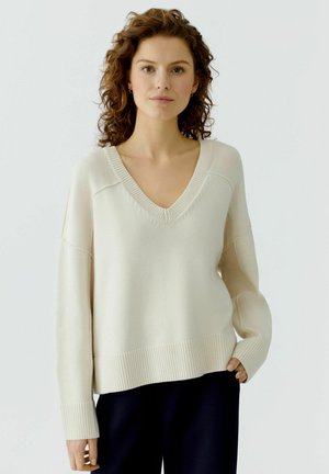 Femme aux cheveux bouclés portant un pull oversize crème à col en V et un pantalon noir, debout devant un fond clair uni.