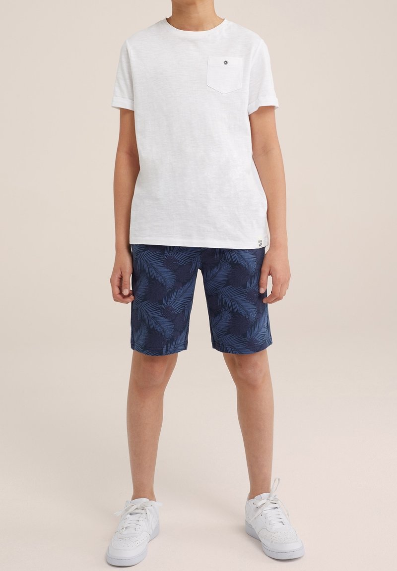 Dziecko ubrane w białą koszulkę z krótkim rękawem i kieszenią na piersi, granatowe szorty w liściasty wzór oraz białe trampki, stojące na beżowym tle.