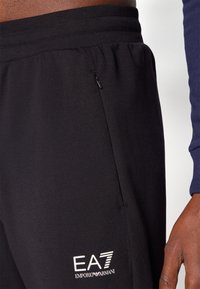 Svarte treningbukser med myk tekstur, elastisk midje, sidelomme med glidelås, og hvit EA7-logo detaljert nederst til venstre.