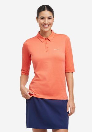 Vrouw die glimlacht en een koraaloranje poloshirt met drie knopen en een donkerblauwe rok draagt, tegen een effen witte achtergrond.
