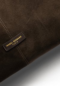 Bruin suede materiaal met een zichtbare textuur, voorzien van een zwart leren label met de opdruk "ISABEL BERNARD" in gouden letters.