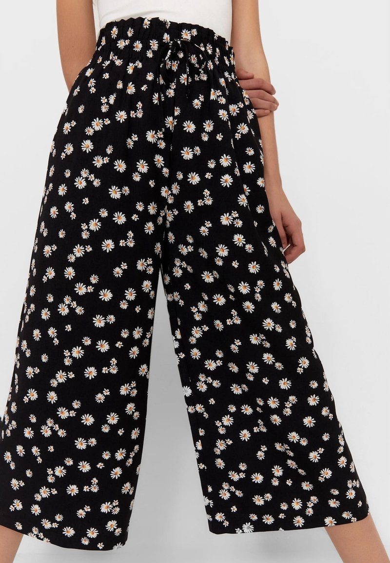 Pantalon noir à jambes larges avec imprimé marguerite blanche et taille élastique, porté avec un haut blanc, montrant le ventre et les jambes.