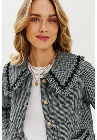 Černobílá károvaná bunda s volánkovým límcem, černými detaily a velkými knoflíky, nošená přes bílý top a vrstvená s barevnými náhrdelníky.