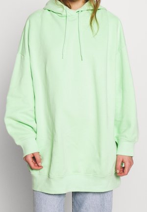 Personne portant un sweat à capuche vert clair oversize avec des cordons de serrage et un jean bleu clair, debout devant un fond uni.