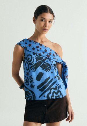 Mujer que lleva una blusa azul de un solo hombro con patrones abstractos y estrellas negras, combinada con una falda negra con botones sobre un fondo liso.