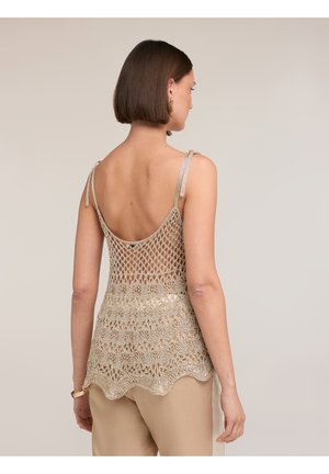 Kvinde med kort brunt hår iført en ærmeløs beige hækletrøje med bølget kant og beige bukser, vist bagfra.