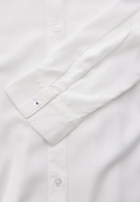 Chemise blanche à manches longues avec une texture lisse, dotée d'un poignet à boutons et d'une subtile touche de violet sur le bouton du poignet.